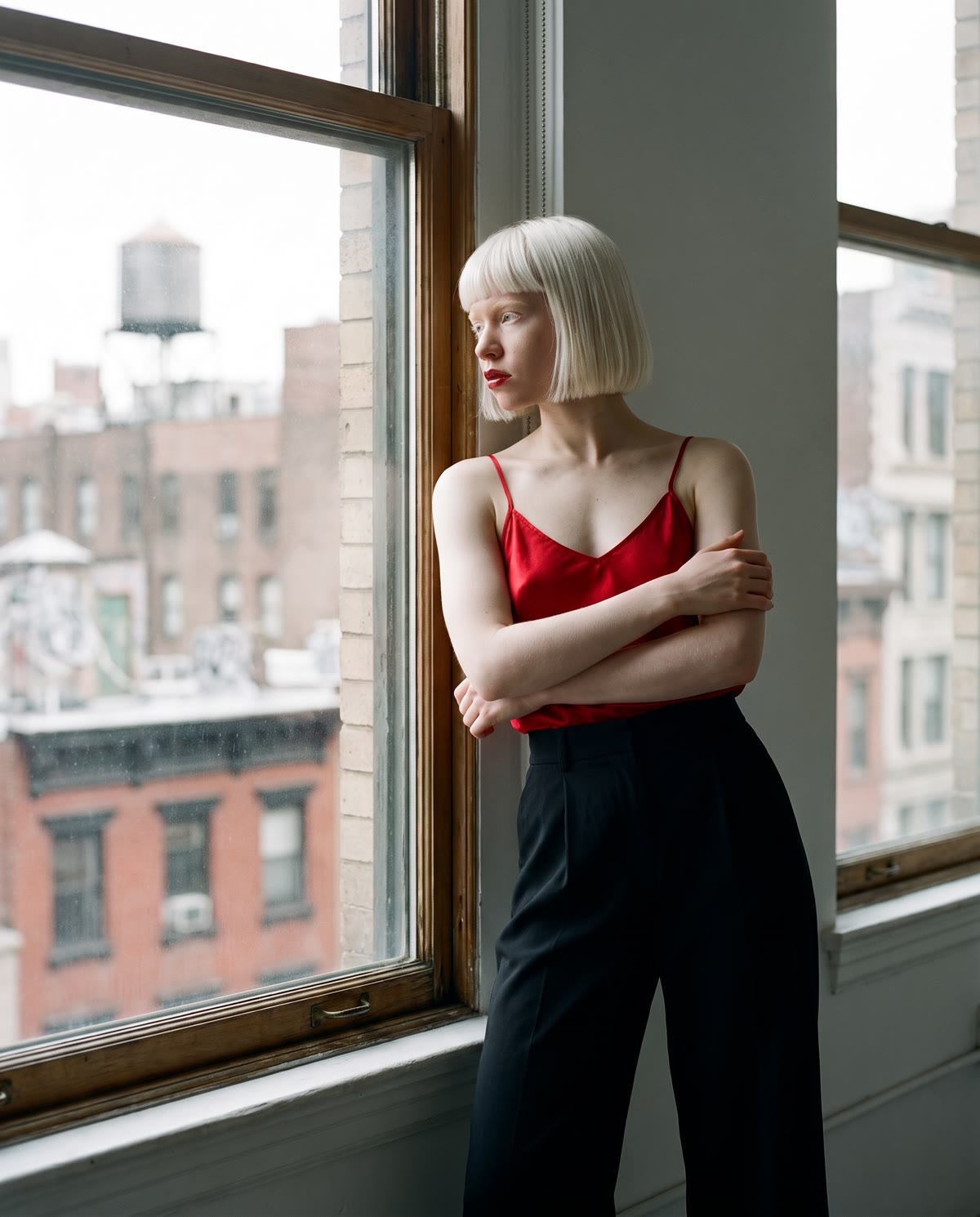 Liv leaning against a tall loft window, arms folded, looking out over Manhattan rooftops